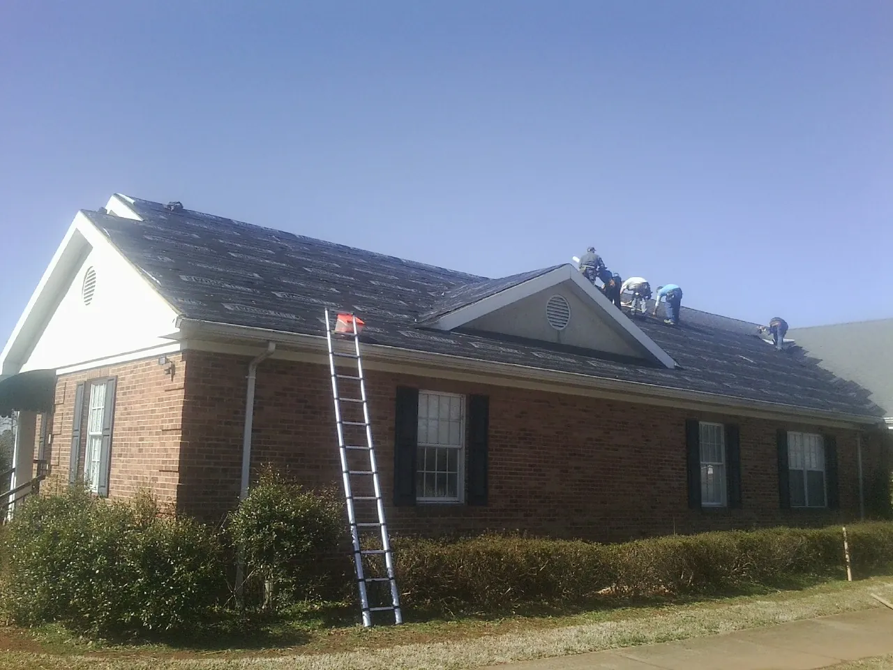 Crew tearing shingles off a brick ranch roof with a ladder set — Metal Roof Repair work by our crew in Noti