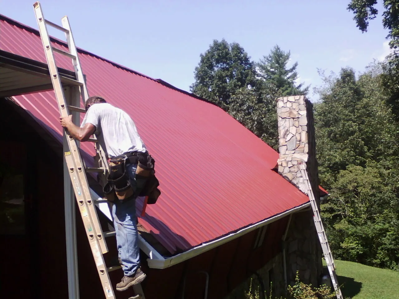 Roofer on a ladder beside a red metal roof and stone chimney — Soffit & Fascia Repair by Roofing Brothers in Noti